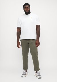 Camiseta blanca de algodón con un pequeño logotipo amarillo en el pecho, acompañada de pantalones verde oliva y zapatillas gris y blancas.