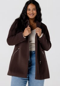 Bruine ritsjas gemaakt van zachte stof, met een getailleerde vorm, metalen ritsen en zijzakken, gedragen over een lichte top en jeans.