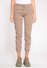 Žena oblečená v béžových slim-fit teplákových kalhotách s elastickými manžetami a odpovídajícími béžovými teniskami, stojící na jednobarevném pozadí.