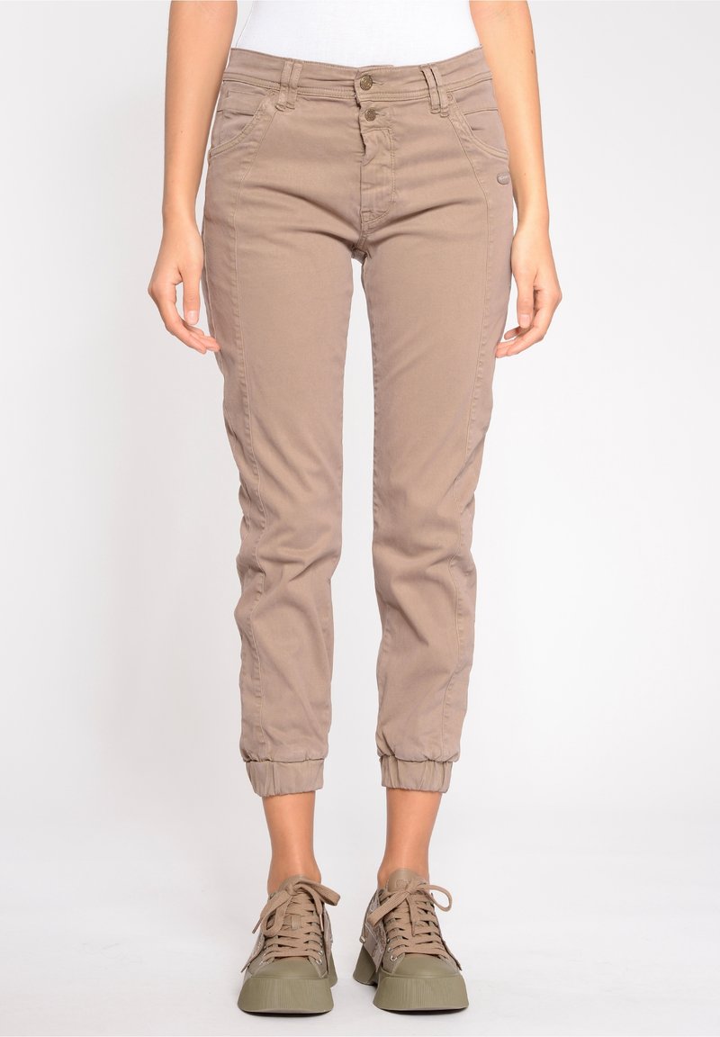 Žena oblečená v béžových slim-fit teplákových kalhotách s elastickými manžetami a odpovídajícími béžovými teniskami, stojící na jednobarevném pozadí.