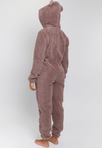Un mono de forro polar marrón, suave y esponjoso, con capucha que presenta pequeñas orejas de oso. Mangas largas y pantalones, la textura parece suave y acogedora. No se aprecian cierres visibles.