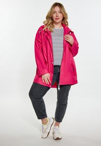 Fényes rózsaszín vízálló dzseki kapucnival, patentgombokkal és zipzárral. Fekete-fehér csíkos pólóra van húzva, sötét farmernadrággal és sportcipőkkel párosítva.