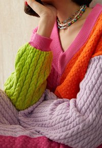 Sweter z kolorowym splotem warkoczowym w zielonych, różowych, pomarańczowych i lawendowych sekcjach. Posiada dekolt w kształcie litery V oraz długie, teksturowane rękawy.