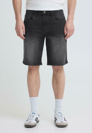 Mann iført falmede svarte denimshorts, hvite sokker og hvite joggesko med svarte striper, stående mot en ensfarget bakgrunn.
