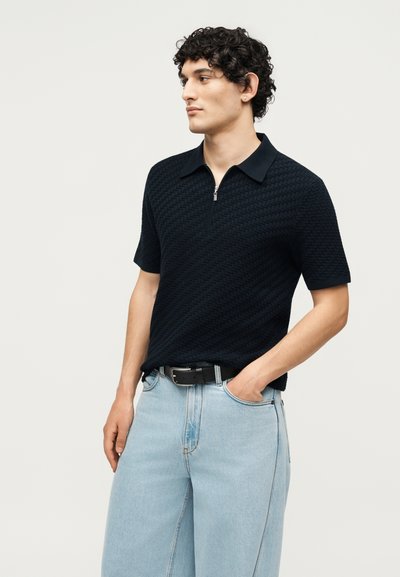 Άνδρας με κατσαρά μαλλιά που φορά κοντομάνικο μαύρο υφασμάτινο πόλο και ανοιχτόμπλε τζιν, στέκεται με το ένα χέρι στην τσέπη.