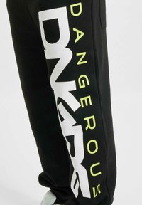 Schwarze Jogginghosen mit einem großen weißen und neon-gelben Grafikdruck mit dem Wort "DANGEROUS" an der Seite. Aus weichem Baumwollmaterial.