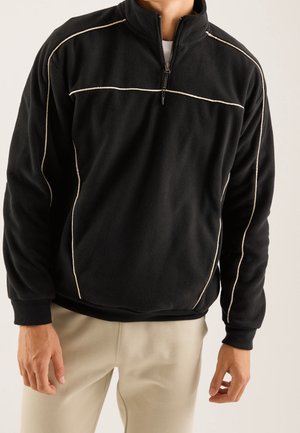 Mann trägt schwarzen Fleece-Pullover mit weißer Paspelierung und halbem Reißverschluss, kombiniert mit hellbeigen Hosen vor einfarbigem Hintergrund.