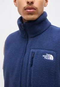 Άνδρας που φοράει μπλε σκούρο φλις γιλέκο με φερμουάρ και ψηλό γιακά, με το λογότυπο "The North Face" στο τσεπάκι στο στήθος.