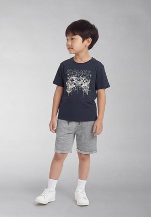 Jonge jongen staand, gekleed in een zwart T-shirt met de tekst "Gamer", grijze korte broek, witte sokken en witte sneakers, kijkt naar links.