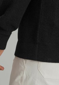 Schwarzer Strickpullover mit strukturiertem Muster, gerippten Bündchen und Säumen sowie einem kleinen Logosticker am unteren Ende. Weiße Hose teilweise sichtbar.