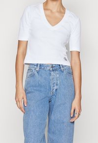 Valkoinen, lyhythihainen puuvillapaita, jossa on V-pääntie. Paidassa on ribattua tekstuuria ja pieni logopatchi vasemmalla puolella hemissä.