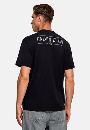 Uomo visto di spalle che indossa una t-shirt nera Calvin Klein e jeans azzurri su uno sfondo chiaro uniforme.