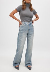 Sieviete stāv ar rokām uz gurniem, ģērbusies pielāgotā pelēkā īsā piedurkņu turtlenekā, gaiši zilās platās biksēs un pelēkos norādītajos apavos.