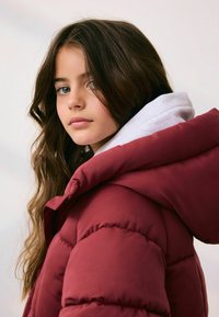 Bordeaux puffer-jakke med en stor hætte, med en glat tekstur og puftede sektioner. Lagd med en lys grå hættetrøje underneath.