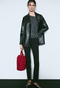 Schwarze Lederblazer über einem gepunkteten Oberteil, kombiniert mit schwarzen Hosen und einer roten Handtasche. Das Model steht selbstbewusst mit Schuhen, die eine Schleifenakzent haben.