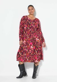 Langärmliges, gemustertes Kleid in Pink, Orange und Schwarz mit einer taillierten Taille und schwingendem Rock. Kombiniert mit schwarzen Cowboy-Stiefeln.