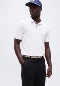 Άνδρας που φοράει λευκό πόλο μπλουζάκι με κοντά μανίκια, μαύρο παντελόνι, μπεζ ζώνη και μπλε καπέλο, στέκεται με το ένα χέρι στην τσέπη, μπροστά σε λευκό φόντο.