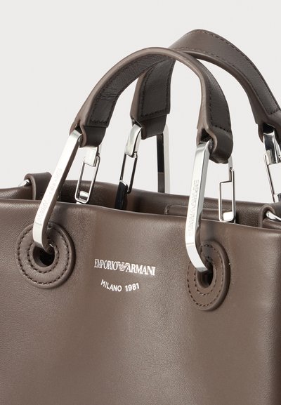 Καφέ δερμάτινη τσάντα Emporio Armani με επενδυμένες λαβές και μεταλλικά εξαρτήματα που παρουσιάζουν το όνομα της μάρκας και το κείμενο "Milano 1981".