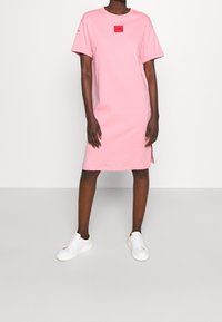 Różowa bawełniana sukienka t-shirtowa z krótkimi rękawami, okrągłym dekoltem, czerwoną łatą z logo, bocznymi rozcięciami i luźnym fasonem, zestawiona z białymi trampkami.