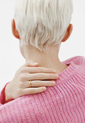 Anillo de oro con un diseño retorcido que se lleva en una mano descansando sobre un suéter de canalé rosa. Cabello corto, texturizado y de color claro visible en el fondo.