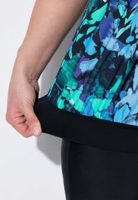 Top colorato con motivo floreale e orlo nero, realizzato in tessuto leggero. Una mano sta tirando l'orlo, accanto a leggings neri.