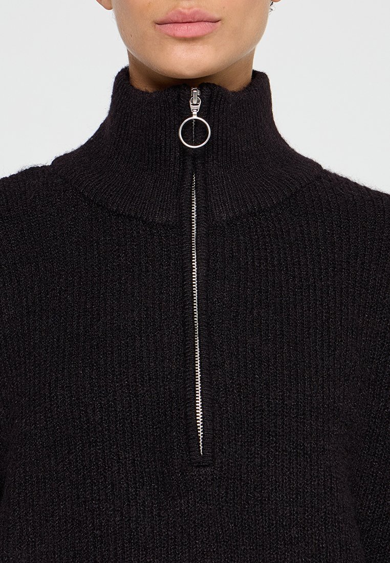 Suéter negro de cuello alto con textura acanalada y cierre de media cremallera con un tirador metálico circular. Suave material de punto que proporciona calidez.