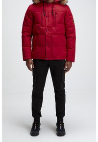Veste doudoune rouge avec une capuche en fourrure, deux poches poitrine et un patch logo. Portée avec un pantalon noir et des baskets noires.