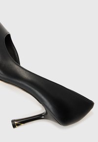 Musta korkokenkä, joka on valmistettu sileästä nahasta, pyöristetyllä kärjellä ja hoikalla stiletto-kannalla, jossa on kultainen yksityiskohta kannan juuressa.