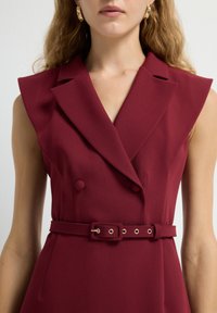 Robe sans manches bordeaux avec un design à double boutonnage, des revers pointus, des épaules structurées et une taille ceinturée avec quincaillerie dorée.