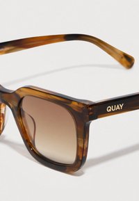 Kilpikonnakuvioiset aurinkolasit, joissa on paksut kehykset, liukuvärjätyt ruskeat linssit ja kullanvärinen "QUAY"-brändäys sankassa, tasainen pinta ja suorakulmainen muoto.