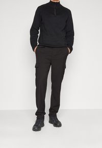 Zwarte fleece sweater met ritssluiting, gecombineerd met zwarte cargo broek met zijzakken. De schoeisel is zwart, met een ruwe textuur en stevige zolen.
