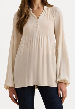 Kvinde iført en cremefarvet plisseret bluse med lange ærmer og knappet halsudskæring samt mørkeblå bukser, stående med armene afslappet ned langs siderne.