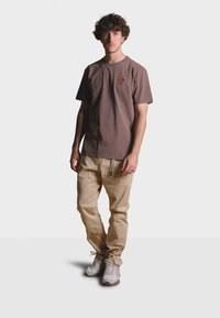 Hnědé bavlněné tričko s krátkým rukávem a malým logem, kombinované se světle béžovými kalhotami a šedými teniskami, stojící na bílém pozadí.