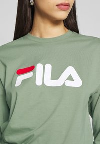 Grüne Baumwollbluse mit langen Ärmeln, die ein auffälliges weißes und rotes FILA-Logo auf der Vorderseite trägt. Sie hat einen Rundhalsausschnitt und eine angenehme, weiche Textur.