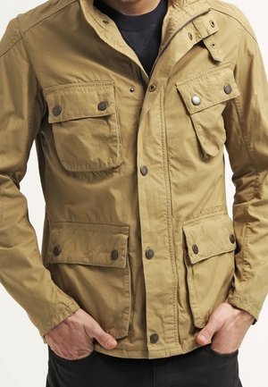 Mann trägt eine beige Freizeitjacke mit vier vorderen Pattentaschen, die mit Metall-Druckknöpfen verschlossen sind, Hände in den unteren Taschen.