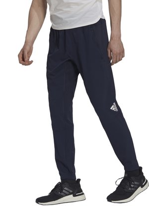 Námořnické modré sportovní kalhoty vyrobené z lehkého materiálu, s elastickým pasem, kapsami na zip a bílým logem Adidas na dolní části nohy.