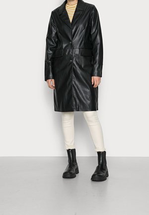 Persoon draagt een lange zwarte leren jas, witte broek, zwarte enkellaarsjes en een geel-wit gestreepte top, staand op een grijze vloer.