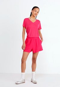 Felle roze V-hals t-shirt gecombineerd met bijpassende high-waisted shorts. Model draagt witte sokken en zilveren ballerina's. Eenvoudig ontwerp; lichtgewicht stof.