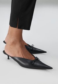 Voeten in zwarte puntige kitten heel mules met kleine strikken, gecombineerd met cropped zwarte broek met zijsplitten.