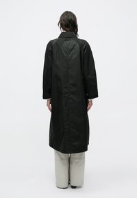 Femeie cu păr închis la culoare, purtând o haină lungă neagră și pantaloni largi de culoare gri deschis, stând cu spatele la fundal alb simplu.