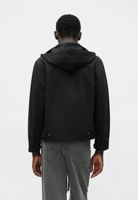 Čierna kapucňová bunda s krátkym strihom, obsahujúca gombíkové detaily na leme a textúrovanú látku. Nosená so sivými džínami.