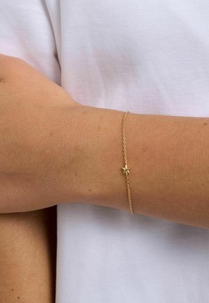 Delikatna złota bransoletka łańcuszkowa z małym zawieszonym gwiazdką, noszona na nadgarstku na tle białej tkaniny.