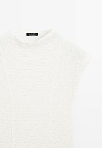 Witte geprofileerde top met korte mouwen en een hoge halslijn. Gemaakt van lichtgewicht stof, met een subtiel kreukpatroon. Merklabel zichtbaar.