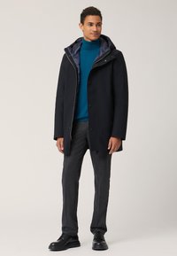 Cappotto in misto lana blu navy con cappuccio, dotato di chiusura con zip e bottoni a scatto. Indossato sopra un dolcevita teal e pantaloni grigi, abbinato a stivali neri.