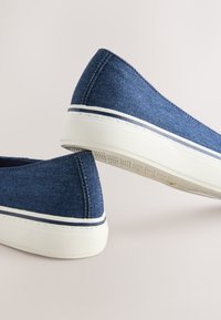 Paire de chaussures slip-on en denim bleu avec semelles en caoutchouc blanches et une bande foncée près du bord de la semelle sur un fond clair.