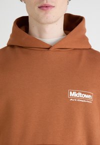 Brązowa bluza z kapturem, wykonana z miękkiego materiału, z białym logo na lewym piersiowym, które brzmi "Midtown Art District."