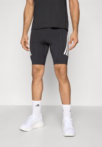 Czarne sportowe spodenki z białymi paskami po bokach, wykonane z gładkiego, elastycznego materiału. Noszone z białymi butami sportowymi i skarpetkami.