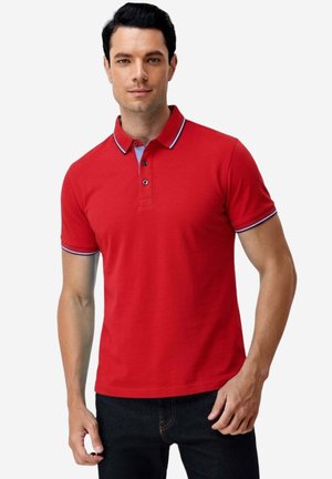 Mann trägt ein rotes kurzärmeliges Poloshirt mit marineblauen und weißen Besätzen an Kragen und Ärmeln, kombiniert mit dunklen Jeans.