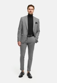 Grå skreddersydd drakt med en teksturert overflate, svart høyhalset genser og lommetørkle. Kombinert med svarte loafers med et strømlinjeformet design.