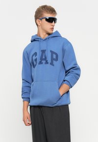 Joven con sudadera azul de GAP, gafas de sol oscuras y pantalones oscuros, de pie con una mano en el bolsillo, mirando hacia un lado.